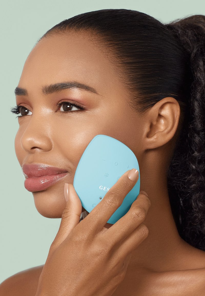 Woman with smooth skin holds a light blue facial cleansing or massage device to her cheek against a pale green background.
