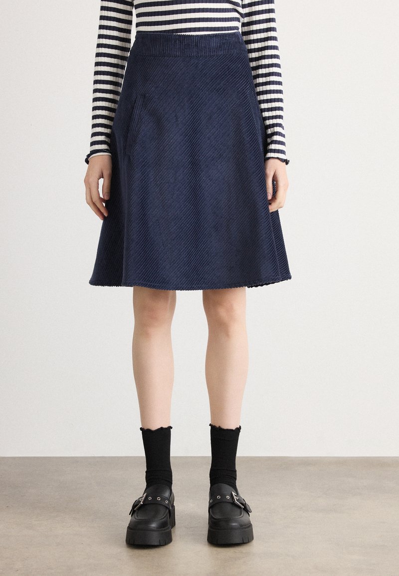 Mörkblå kordflodshort A-linjeformad kjol med sidofickor, ribbad textur och en fåll som försiktigt vidgar sig. Kombineras med en randig långärmad topp och svarta skor.