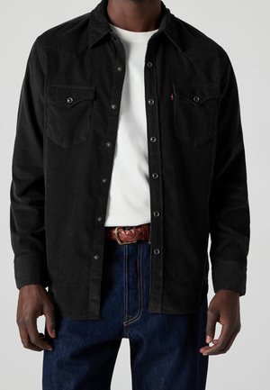 Homme portant une chemise en velours côtelé noire boutonnée sur un t-shirt blanc, un jean bleu et une ceinture en cuir marron tressée, visible du cou aux hanches.