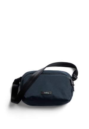 Sac bandoulière bleu marine en tissu ; doté d'une fermeture éclair, d'une bandoulière réglable et d'un écusson logo noir à l'avant. Texture lisse.