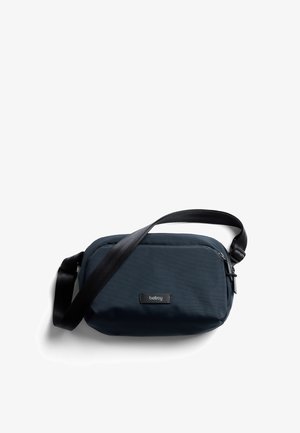 Sac bandoulière bleu marine en tissu ; doté d'une fermeture éclair, d'une bandoulière réglable et d'un écusson logo noir à l'avant. Texture lisse.
