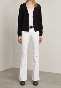 Cardigan noir boutonné par-dessus un débardeur blanc, associé à un jean évasé blanc à taille haute et des bottines noires à talons pointus.