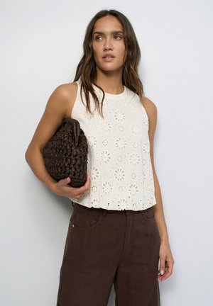 Femme portant un haut blanc sans manches à œillets et un pantalon marron, tenant une grande pochette tressée marron foncé sur un fond clair uni.