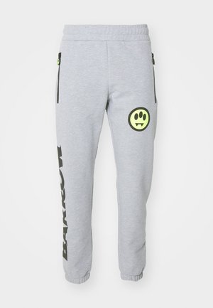 Pantalones de chándal grises con bolsillos laterales con cremallera, presentan un diseño de texto vertical negro y un logo gráfico redondo verde en el muslo izquierdo.