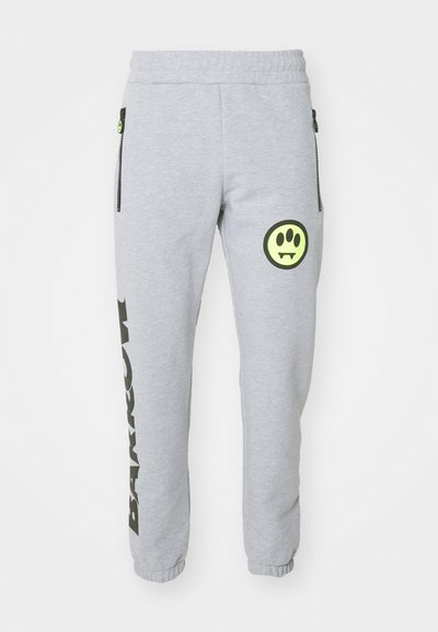 Pelēki sporta bikses ar rāvējspējamiem sānu kabatām, ar melnu vertikālu teksta dizainu un apaļu, zaļu grafisko logotipu kreisajā augšstilbā.