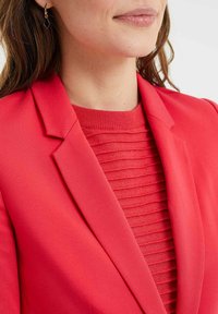 Röd blazer med en skarp slag, lager på lager över en texturerad ribbad topp i en matchande nyans. Modellen bär en geometrisk örhänge.