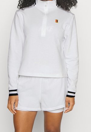 Personne portant un haut blanc à manches longues avec fermeture éclair et poignets rayés noirs, ainsi qu’un short blanc assorti, debout devant un fond uni.