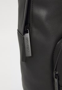 Zaino nero con una texture liscia simile al cuoio, chiusura con zip e tiranti in metallo marchiati, dal design elegante e moderno.