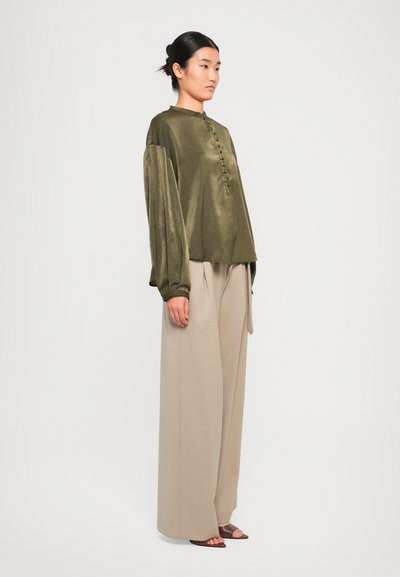 Kvinne som står i beige vidbukser, olivengrønn langermet bluse med knapper, og brune åpne hæler mot en ensfarget hvit bakgrunn.