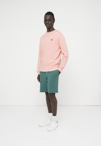 Ανοιχτό ροζ φούτερ, πράσινο σορτς, λευκά αθλητικά παπούτσια. Κεντημένο λογότυπο στο στήθος και στα σορτς. Λεπτή υφή, άνετη εφαρμογή, απλός σχεδιασμός.