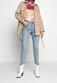 Veste en velours côtelé beige avec poches, t-shirt multicolore en tie-dye, jean skinny bleu clair, et bottes de cowboy blanches à talons.