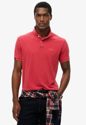 Rød polo shirt med krave og to knapper, med et diskret logo. Sammen med mørke jeans og en ternet skjorte bundet om livet.