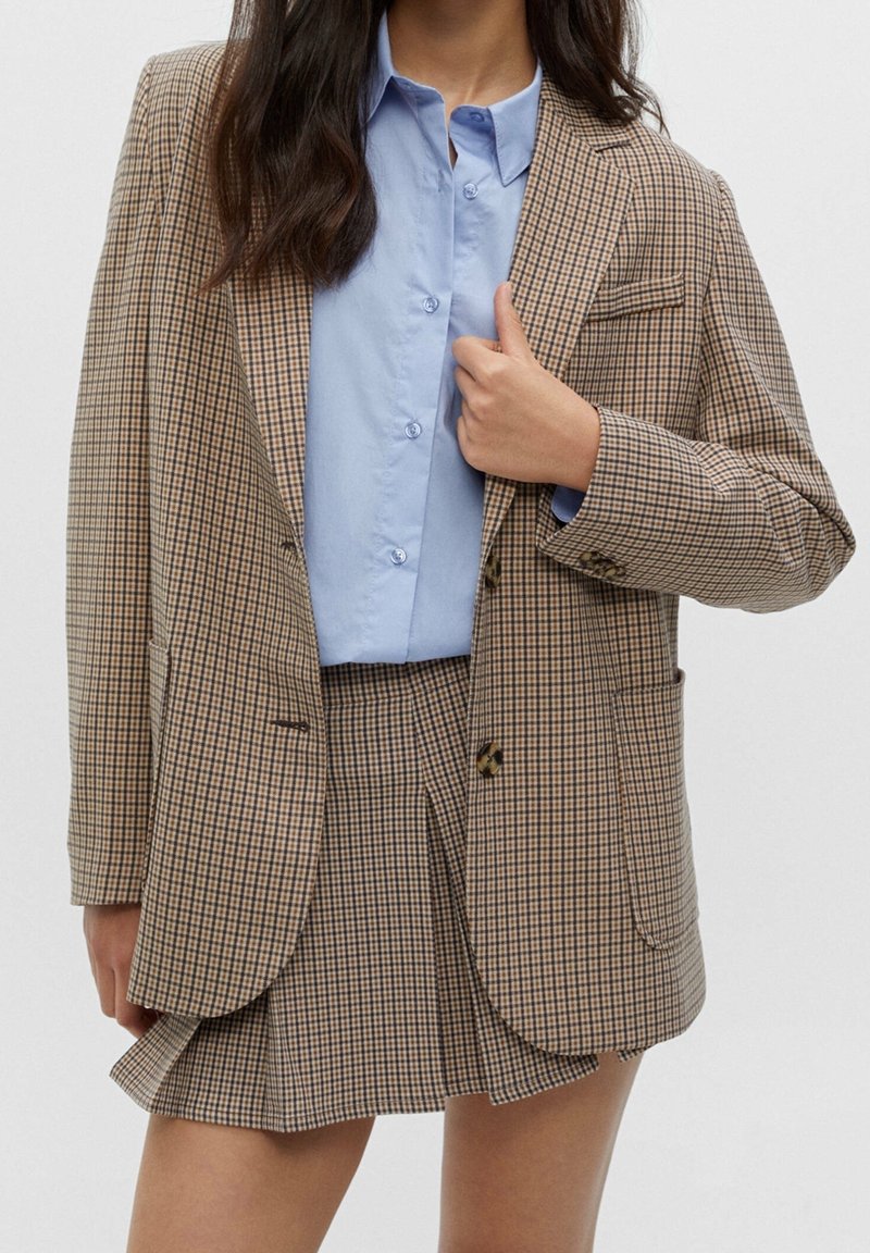 Femme portant un blazer à carreaux beige et une jupe plissée assortie avec une chemise bleu clair boutonnée, tenant le revers du blazer.