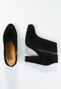 Botas de tobillo de ante negro con tacón alto, que cuentan con una cremallera lateral y una punta redondeada. El tacón tiene un acento brillante.