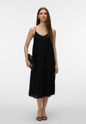 Mujer con cabello largo y castaño, vestida con un vestido midi negro sin mangas, sosteniendo un bolso de mano negro y usando sandalias marrones con tiras.