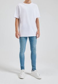 Bílá bavlněná tričko s krátkým rukávem, kombinované se světle modrými skinny džíny. Dokončeno s bílými teniskami a jednobarevným pozadím.