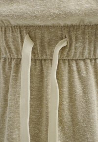 Ceinture en tissu beige avec élastique et deux cordons crème pendant à l’avant.