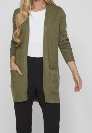 Femme portant un long cardigan vert olive ouvert à l'avant avec des poches, un haut blanc et un pantalon noir sur un fond gris uni.