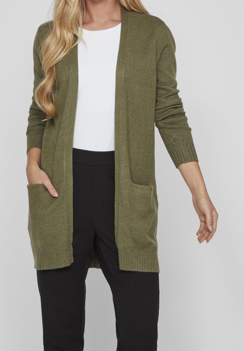 Femme portant un long cardigan vert olive ouvert à l'avant avec des poches, un haut blanc et un pantalon noir sur un fond gris uni.