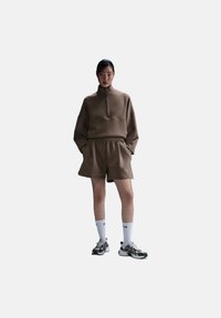 Sudadera de forro polar marrón con cuello de media cremallera combinada con pantalones cortos a juego. El modelo lleva calcetines blancos y zapatillas deportivas grises. Diseño simple, ajuste casual.