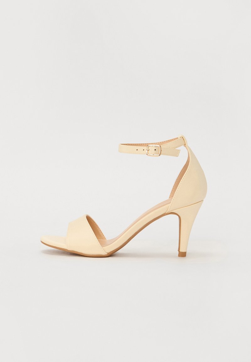 Beige high-heeled sandal with a closed back, open toe, ankle strap with buckle, smooth material, and tapered heel design.