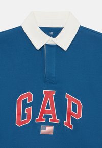 Plava polo majica s bijelim ovratnikom, s velikim crvenim "GAP" logotipom i malim detaljem američke zastave. Pamuk s glatkom teksturom.