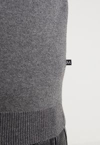 Grå stickad tröja med en texturerad yta, med en ribbad nederkant och en liten svart etikett p å sidan.