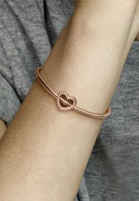 Bracciale in oro rosa con design intrecciato, dotato di un fermaglio a forma di cuore decorato con piccole perline. Texture liscia sul polso.