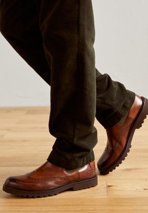 Braune Leder-Schuhe mit Brogue-Details, Gummisohle und runder Spitze. Kombiniert mit dunkelgrünen Cordhosen, die einen strukturierten Stoff zeigen.