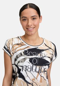 Short-sleeve, printed T-shirt featuring abstract cream, beige, and black patterns. Text reads "Beauty of nature" in sequins. Lightweight fabric.