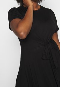 Robe noire à manches courtes avec un détail de nœud à la taille, présentant une texture douce et une coupe décontractée. Design simple et classique.