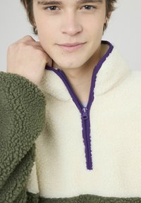 Pull-over en polaire crème et verte à demi-zip avec une fermeture éclair violette. Texture douce, col haut et blocs de couleur contrastante.