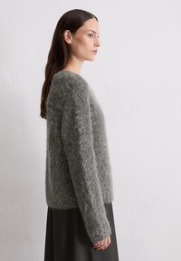 Puszysty szary sweter dzianinowy o luźnym kroju, długich rękawach i okrągłym dekolcie, zestawiony z ciemną spódnicą. Gładka faktura i miękki wygląd.