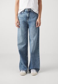 Personne portant un jean en denim bleu à jambes larges, des baskets blanches et un t-shirt blanc sur un fond clair uni.