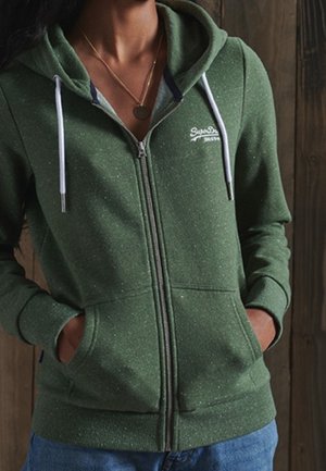 Woman wearing a green zip-up hoodie with white drawstrings and hands in pockets, against a wooden background.