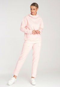 Różowy dres z kapturem o luźnym kroju i długich rękawach, w zestawie z pasującymi różowymi spodniami typu straight-leg. Obie części charakteryzują się gładką fakturą i swobodnym stylem.