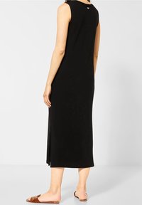 Kvinna som bär en ärmlös svart midiklänning med en sidoflik, kombinerad med bruna platta sandaler, stående mot en vit bakgrund.