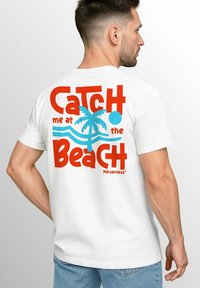 White short-sleeve T-shirt featuring a colorful graphic on the back with red and blue text reading "Catch me at the Beach" and a palm tree design.