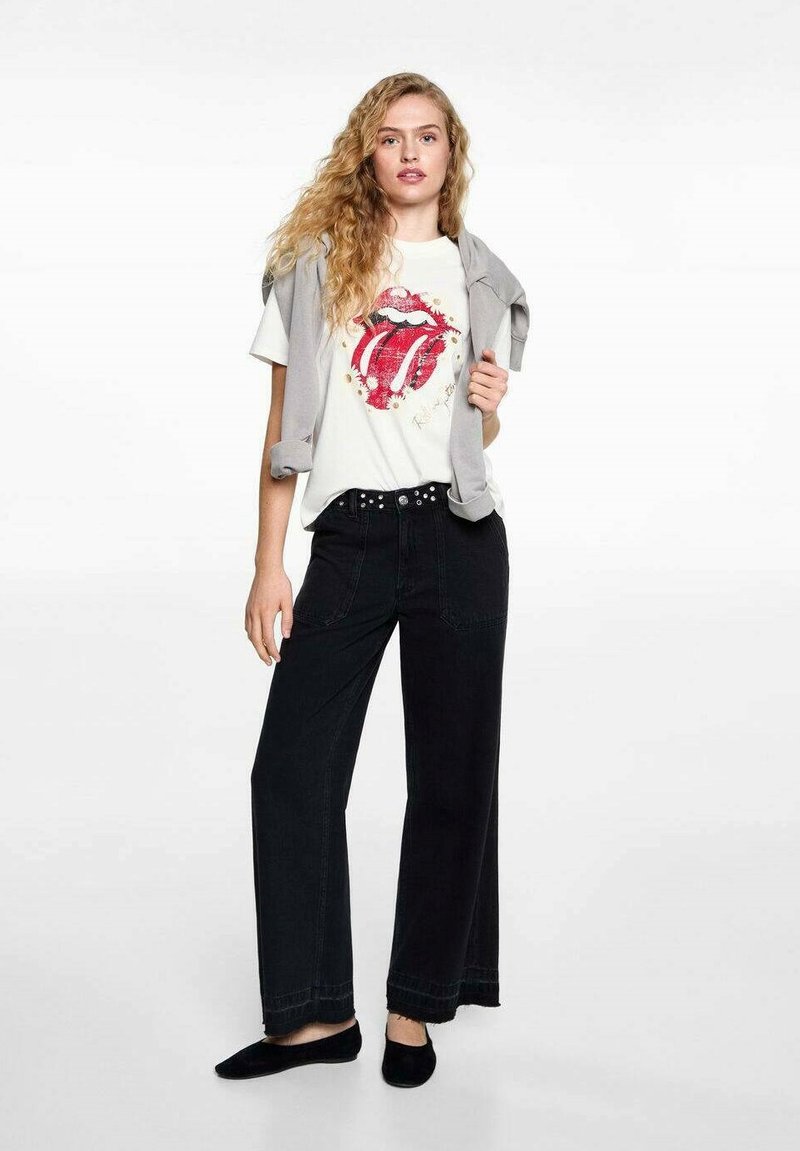 Grafická tričko s červeným logom Rolling Stones, ktorým sú doplnené čierne široké nohavice s vysokým pásom a svetlo šedý sveter zviazaný na pleciach.