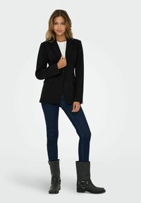 Černý blazer s přiléhavým střihem, klopovými límci a dvěma předními kapsami, v kombinaci s tmavými úzkými džíny a černými vysokými botami s kovovými akcenty.