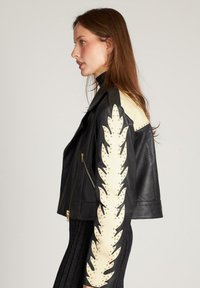 Vrouw in een zwarte leren jas met crèmekleurig vlamvormig patroon en gouden studs op de mouw, gecombineerd met een zwart geribd outfit, naar links gekeerd.