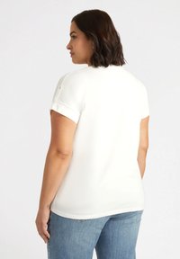 Top blanco de manga corta con una textura suave y escote redondeado, combinado con jeans de color azul claro, visto desde la parte de atrás.