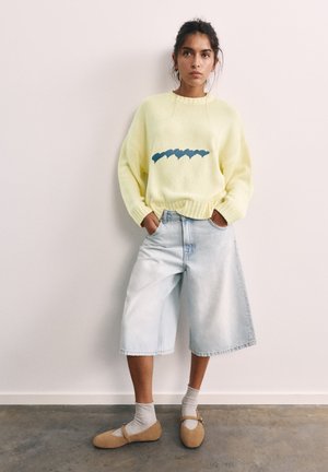 Frau steht mit den Händen in den Taschen, trägt einen gelben Pullover mit blauem Design, helle weite Denim-Shorts, graue Socken und hellbraune Schuhe vor einer weißen Wand.