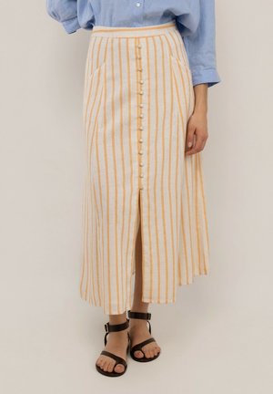 Mujer con falda midi de botones en la parte delantera con rayas verticales en beige y naranja y camisa azul de manga larga, con sandalias negras de tiras en el tobillo.