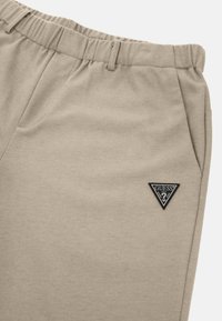 Bežinės sportinės kelnės su elastinga juosta, su šonine kišene ir trikampio formos juodu Guess logotipo ženklu. Lygus medvilnės audinio paviršius.