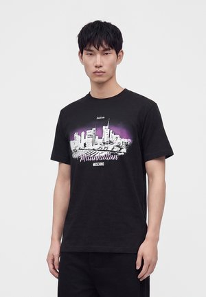 Jeune homme aux cheveux courts et foncés portant un t-shirt noir avec un graphisme de ligne d'horizon de ville en blanc et violet et texte "Mlanhattan Moschino."