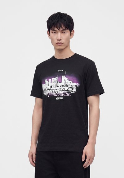 Jeune homme aux cheveux courts et foncés portant un t-shirt noir avec un graphisme de ligne d'horizon de ville en blanc et violet et texte "Mlanhattan Moschino."