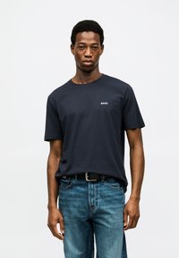 Άνδρας που φοράει μπλε σκούρο t-shirt BOSS, μπλουζάκι μπουσουλαρισμένο σε μπλε τζιν με μαύρη ζώνη, στέκεται μπροστά σε ένα απλό λευκό φόντο.