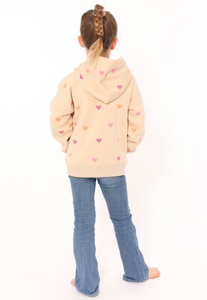 Beige Kapuzenpullover mit mehrfarbigen Herzen in Pink und Orange, ausgestattet mit einer großen Kapuze und geripptem Saum. Kombiniert mit blauen Schlagjeans.