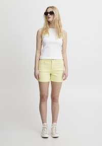Top de tirantes blanco con textura, pantalones cortos amarillos y zapatillas blancas. Los pantalones cortos son de mezclilla con un ajuste clásico. El modelo lleva gafas de sol negras.